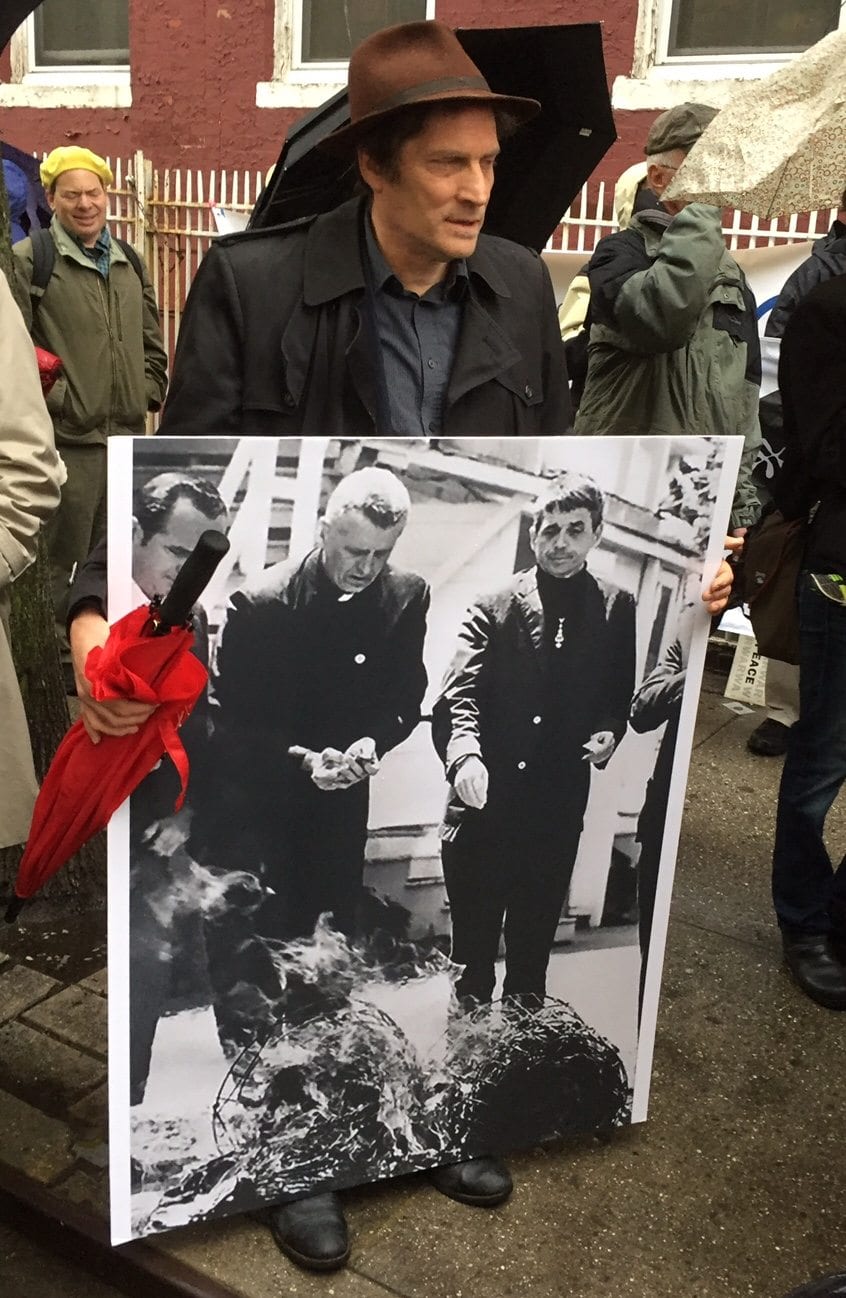 A mourner carries a photo of priests Phil and Dan Berrigan burning draft records during a 1968 protest of the Vietnam War. (Photo by RNS/Heidi Thompson.)