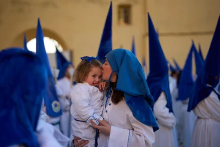 A hooded penitent of the Nuestro Padre Jesus Cautivo y Nuestra Senora de la Estrella brotherhood kisses her child during a Holy Week procession in Dona Mencia, southern Spain, Tuesday, March 31, 2026. (Credit: Manu Fernandez/AP.)