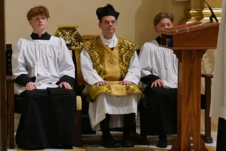 Father John Ubel, pastor of the Church of Saint Agnes, wears the traditional biretta hat and vestments as he sits between altar servers during one of the Catholic parish’s Latin Masses in St. Paul, Minn., Sunday, June 28, 2026. (Credit: Giovanna Dell’Orto/AP.)
