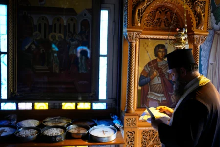 Father Nikolaos Vlavianos blesses the parishioners' Fanouropites, orange-flavored cakes, as an icon of Saint Fanourios, known in Greek as "The Revealer," is seen at right at the Church of St. Fanourios in Athens, Tuesday, Aug. 25, 2026. (Credit: Thanassis Stavrakis/AP.)