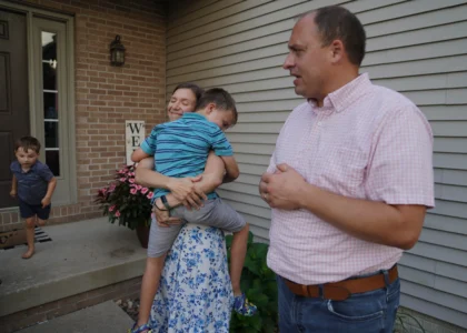 Lizzy Taggart holds her son Fulton, 8, while standing with her husband, Joe, outside their home in Bloomington, Ill., on July 16, 2026. (Credit: Jessie Wardarski/AP.)